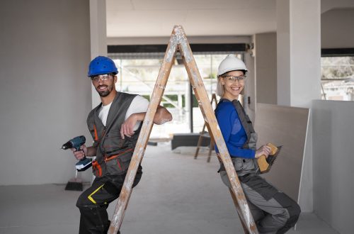 man and woman wearing hard hats standing with a ladder between them inside a room that is under renovation